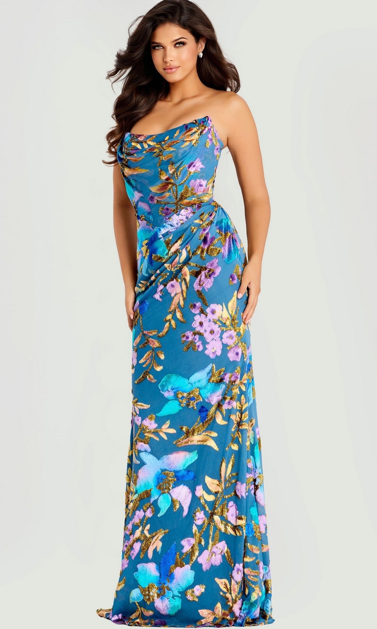 A woman models the Jovani 47654, a fitted, floor-length blue prom dress with a vibrant floral and bird pattern, standing against a plain background.